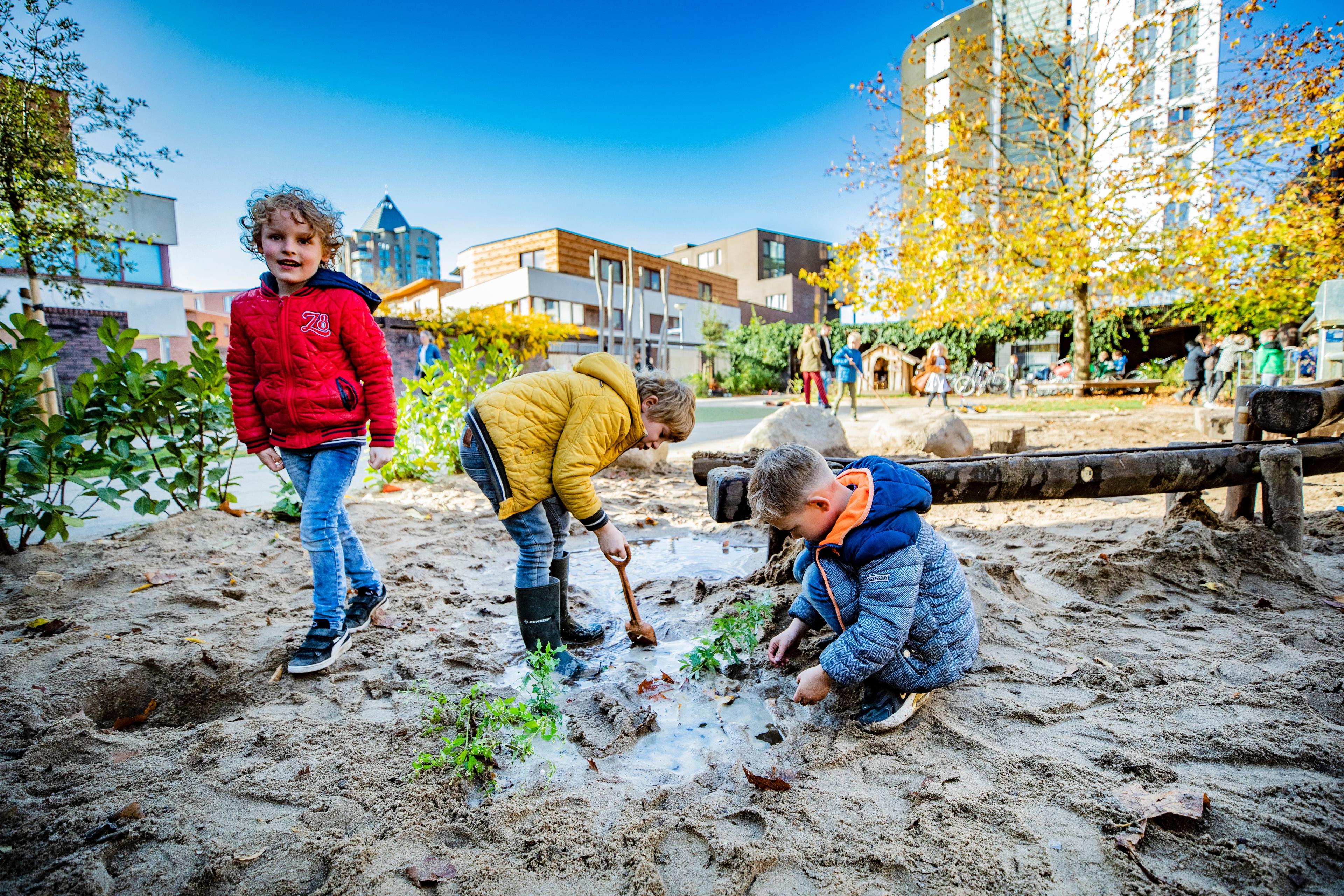 3 kinderen spelen op de voorgrond in de modder met gebouwen op de achtergrond